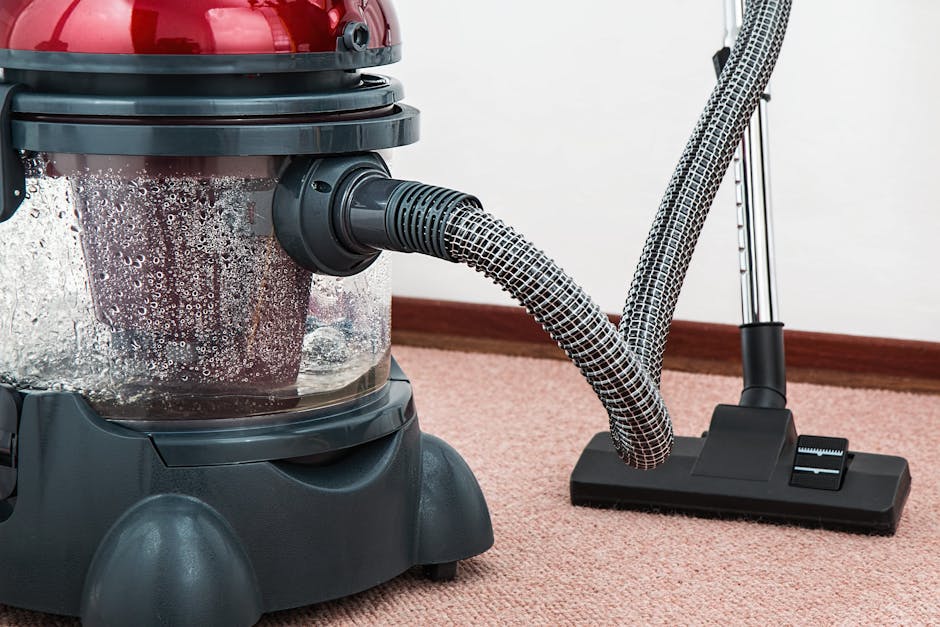 A professional cleaner dressed in a white protective suit, mask, and gloves performing carpet steam cleaning in a modern living room. The individual is operating a vacuum-like machine that releases st